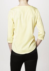 Blouse jaune à manches trois-quarts avec un col rond et un ourlet arrondi. Textura lisse, associée à un pantalon noir, vue de dos.