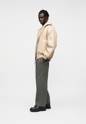 Άνδρας που στέκεται πλευρικά φορώντας μπεζ μπουφάν με κουκούλα, γκρι παντελόνι με φαρδιά πόδια και μαύρα χοντρά παπούτσια σε λευκό φόντο.