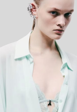 Giovane donna con capelli tirati indietro indossa una camicia trasparente verde menta sopra un bralette in pizzo e un elaborato orecchino con cristalli all'orecchio sinistro.