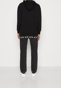 Černá mikina s kapucí ve volném střihu, s bílým logem na lemu, spárovaná s tmavě šedými kalhotami a bílými teniskami.