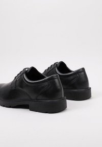 Une paire de chaussures de ville en cuir noir à lacets avec des semelles en caoutchouc épaisses, vues de dos sur un fond blanc.