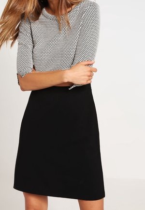 Femme portant une jupe noire et un haut à manches trois-quarts à motifs, debout les bras croisés devant un fond uni.