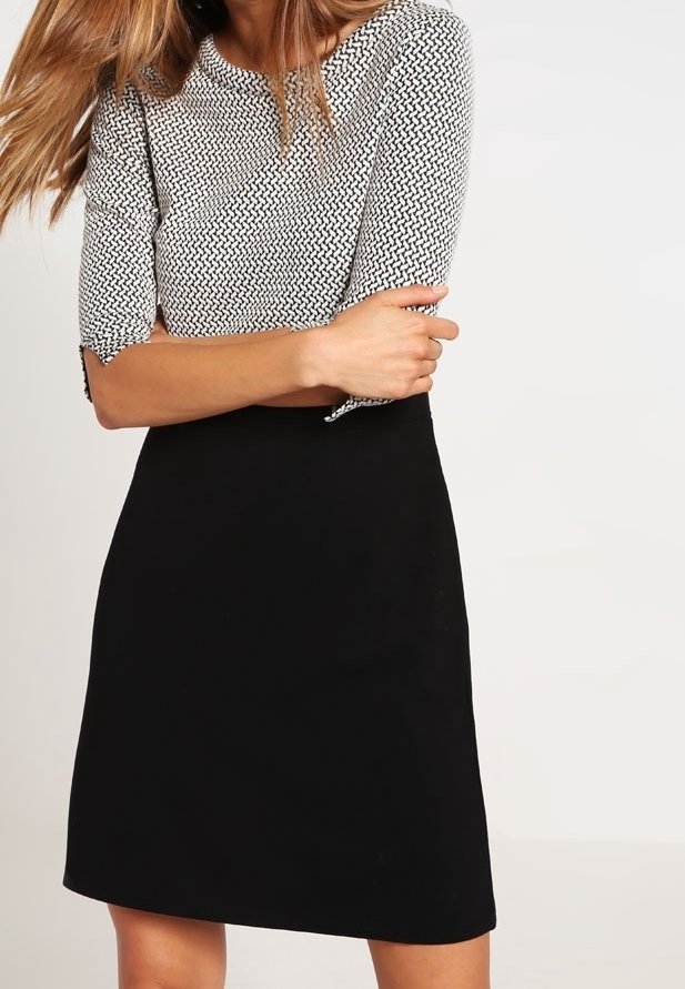 Femme portant une jupe noire et un haut à manches trois-quarts à motifs, debout les bras croisés devant un fond uni.