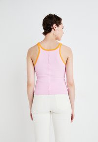Roze ribgebreide tanktop met oranje afwerking, voorzien van een aansluitend ontwerp en een knoopsluiting aan de achterkant. Gecombineerd met witte broek.