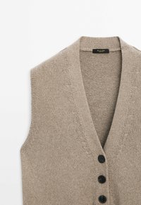 Beige neuleliivi, jossa on V-pääntie, viisi mustaa nappia ja hienovaraiset ommeltiedot kauluksessa ja käsivarsikoloissa.