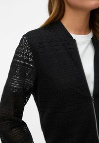 Zwarte kanten bomberjack met ritssluiting, voorzien van textuurpatronen en een geribde kraag, met ingewikkelde uitsnijdingen.