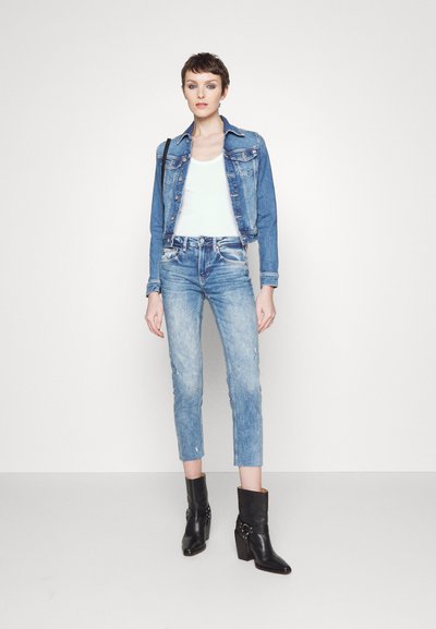 Femme portant une veste en denim bleu et un jean, un débardeur blanc, des bottines noires, debout contre un mur blanc.