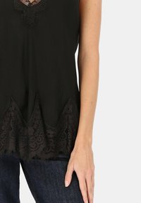 Czarna, bezrękawnikowa bluzka z koronkową lamówką o falistym brzegu i misternych wzorach, wykonana z lekkiego materiału.