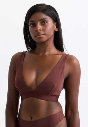 Junge Frau mit dunkler Haut und langen Haaren, die ein braunes ärmelloses Crop-Top und passende Unterteile vor einem einfarbigen Hintergrund trägt.