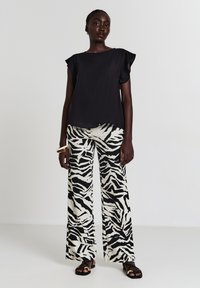 Donna che indossa una maglietta nera larga, pantaloni a gamba larga con stampa zebra, sandali neri con anelli dorati e braccialetti dorati su uno sfondo grigio semplice.