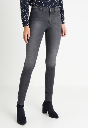 Frau trägt ausgewaschene schwarze Skinny Jeans, schwarze Stiefeletten und eine marineblaue Bluse mit weißen Punkten, steht vor weißem Hintergrund.