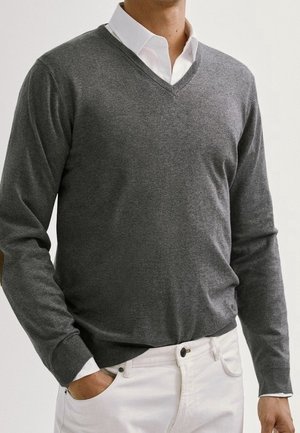 Mann trägt einen grauen V-Ausschnitt-Pullover über einem weißen Hemd mit Kragen, kombiniert mit weißen Hosen, steht mit einer Hand in der Tasche.