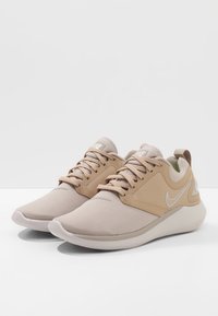 Beige träningsskor med en slät textilöverdel, räfflade detaljer och en vadderad sula. Har en vit Nike-logotyp på sidan.