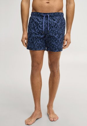 Mężczyzna noszący granatowe kąpielówki z jasnoniebieskim abstrakcyjnym wzorem i sznurkiem, stojący boso na jednolitym tle.