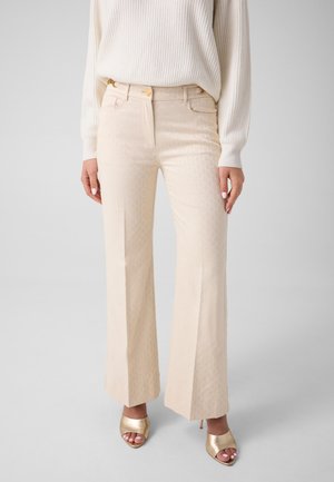 Vrouw draagt beige, hooggetailleerde, wijde broek met patroon, witte geribde trui en gouden open-toe hoge hakken tegen een effen achtergrond.