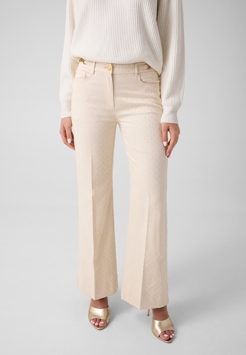 Femme portant un pantalon large beige à motifs taille haute, un pull blanc côtelé et des talons ouverts dorés sur un fond uni.
