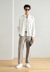Camicia bianca a maniche lunghe, pantaloni grigi e sneakers bianche. Tiene in mano un tablet nero. Capelli corti e ricci e occhiali da sole. Stile semplice e moderno.