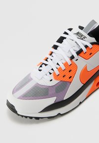 Nike Air Max lenkkarit, joissa on valkoinen, harmaa ja musta yläosa. Varustettu oransseilla yksityiskohdilla, kuviollisella purppurakankaalla ja tekstuurilla varustetulla pohjalla. Valkoiset nauhat.