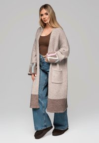 Cardigan grigio chiaro lavorato a maglia con maniche a motivi rosa e blu, abbinato a jeans blu a vita alta e pantofole nere.
