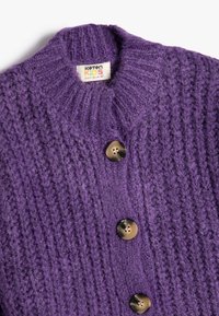 Lila gestrickte Strickjacke für Kinder mit hohem Kragen, versehen mit großen runden braunen Knöpfen und einem strukturierten Stichmuster.