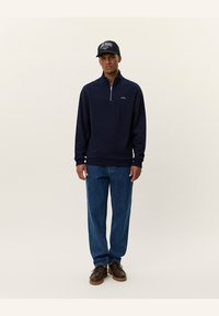 Navy sweatshirt with a zip collar, light ribbed cuffs, and hem; paired with blue denim jeans and dark brown shoes; model wears a black cap.