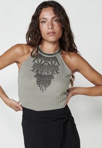 Žebrovaný halter top v světle šedé barvě s černým korálkovým vyšíváním tvořícím geometrický vzor kolem výstřihu. Nosený přes černou sukni.