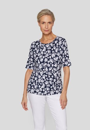 Frau in einem marineblauen, kurzärmeligen Oberteil mit weißen Blumenmustern und weißen Hosen, die vor einem schlichten grauen Hintergrund steht.