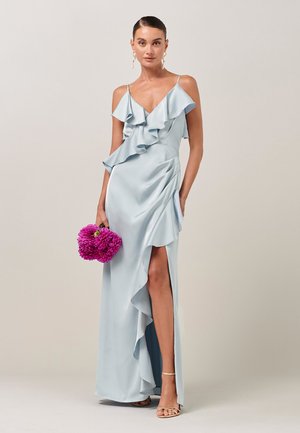 Kvinde i lyseblå satin kjole med flæser, spaghetti-stropper og lårslids, holder lyserøde blomster, står foran en neutral baggrund.