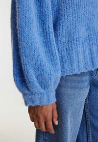 Gros plan sur une main portant un pull en tricot bleu clair avec des manches bouffantes, associé à un jean en denim bleu.