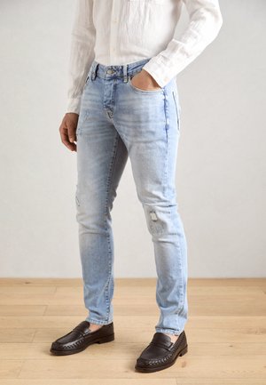 Light blue denim jeans with a slim fit, featuring distressed patches and a classic five-pocket design. Paired with dark loafers.