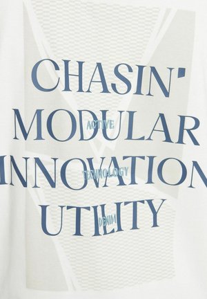 Vitt tyg med djärv blå text som stavar "CHASIN' MODULAR INNOVATION UTILITY ACTIVE TECHNOLOGY DENIM", med ett lager av grafiskt mönster.