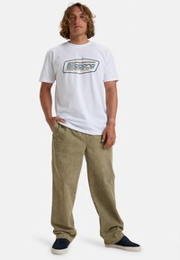 Tricou alb cu logo "Billabong" albastru și galben, asortat cu pantaloni verde măsliniu. Pantalonii sunt largi, confecționați dintr-un material moale.