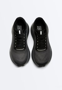 Paire de baskets athlétiques noires et gris foncé avec lacets, dessus en maille, et semelles épaisses et rembourrées vues de dessus sur un fond clair.