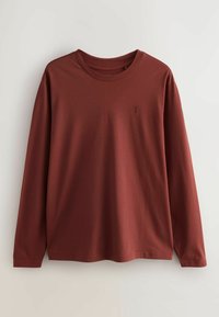 Ilgi piedurkņu, rūsu krāsas krekls, izgatavots no gludas kokvilnas auduma. Tam ir apaļa kakla izgriezuma forma un neliela logotipa emblēma krūtīs.