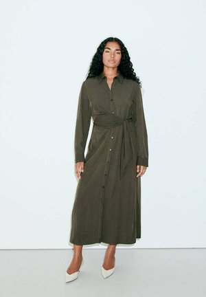 Femme aux longs cheveux bouclés portant une robe longue boutonnée vert olive avec une ceinture à la taille, debout devant un fond clair uni, chaussée de talons blancs.
