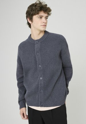 Cardigan en tricot gris avec un col rond. Présente une texture côtes, une fermeture à six boutons et des manches longues. Porté sur une chemise de couleur claire.