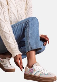 Personne accroupie portant un jean bleu, un pull crème en tricot torsadé et des baskets blanches à plateforme avec des motifs abstraits colorés.