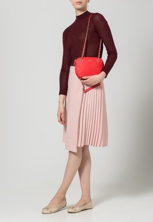 Donna con top a maniche lunghe a coste color bordeaux, gonna plissettata rosa, ballerine beige, che tiene in mano una piccola borsa a tracolla rossa con catena dorata.