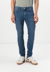 Una persona está de pie vistiendo unos pantalones vaqueros ajustados azules y zapatillas blancas, acompañados de una camiseta gris, frente a un fondo blanco liso.