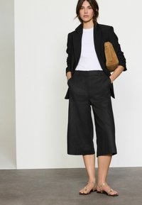 Blazer negro ajustado sobre una camiseta blanca, combinado con culottes negros y sandalias planas metálicas. Bolsa de ante marrón con un asa superior.