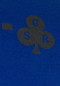 Modrá látka s černým motivem symbolu klub s písmeny G, R a S uvnitř tvaru. Textura je hladká a jemná.