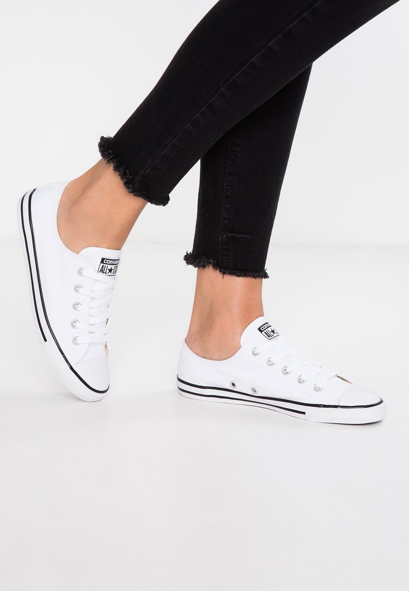 White canvas sneakers with black accents and rubber sole. Features lace-up design and a rounded toe. Jeans have frayed hem.