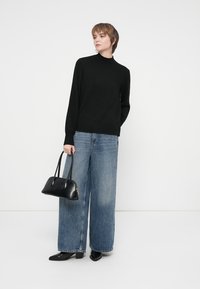 Νεαρός/ή με μαύρο πουλόβερ τζάκετ, φαρδιά μπλε τζιν παντελόνια, μαύρες γόβες και κρατώντας μια μαύρη τσάντα μπροστά σε απλή φόντο.