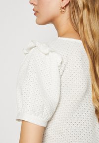 Blusa blanca con mangas abullonadas, que presenta un patrón texturizado, detalle de escote con volantes y un diseño transpirable.