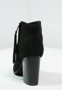Botín ankle de ante negro con tacón afilado, que cuenta con una cremallera lateral y borlas decorativas. Tacón negro suave con un acabado mate.