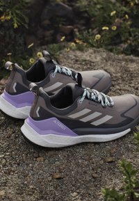 Para szaro-fioletowych butów do biegania Adidas Terrex na żwirze, w tle małe żółte kwiaty.