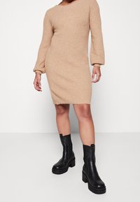 Robe en tricot côtelé beige avec de longues manches bouffantes, un col rond et une coupe à mi-cuisse, assortie à des bottines noires à semelle épaisse.