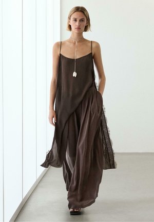 Jeune femme marchant à l'intérieur, portant une robe fluide marron foncé à fines bretelles et un long collier argenté avec des pendentifs en forme de feuilles.