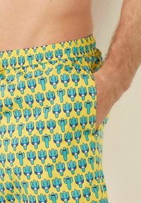 Pantaloni da nuoto realizzati in tessuto leggero, caratterizzati da uno sfondo giallo vivace con un motivo a cactus ripetuto in blu e verde.