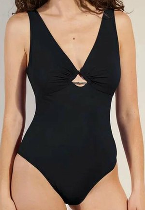 Donna che indossa un costume intero nero con spalline larghe e un piccolo dettaglio con taglio frontale sotto il seno.